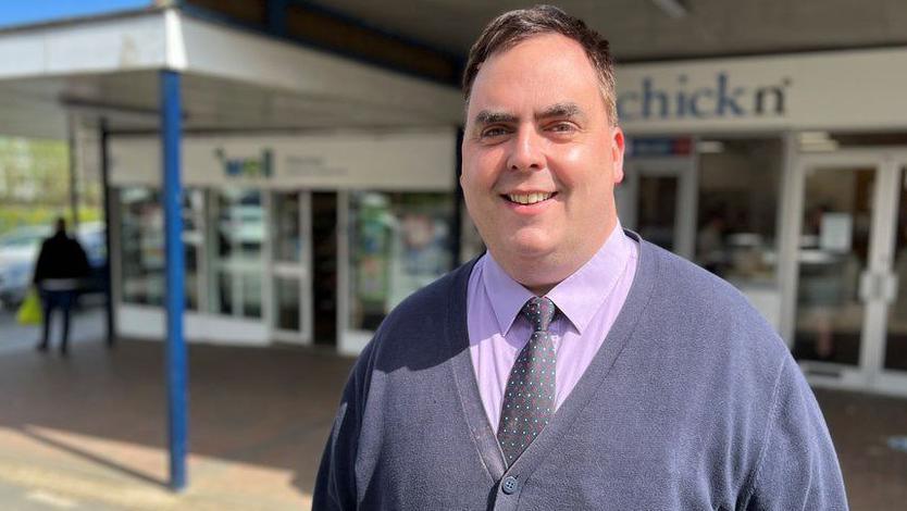 Kerry Smith wearing a cardigan, purple shirt and spotted tie. His hair is receding. He is smiling. He is standing outside in front of a parade of shops