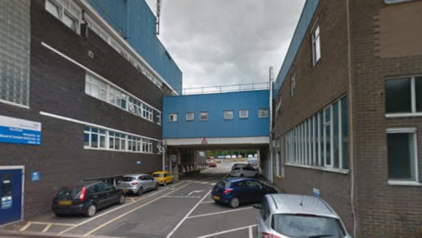 A hospital building - two brick blocks with a small road running between them are joined by a bridge, which is painted blue.