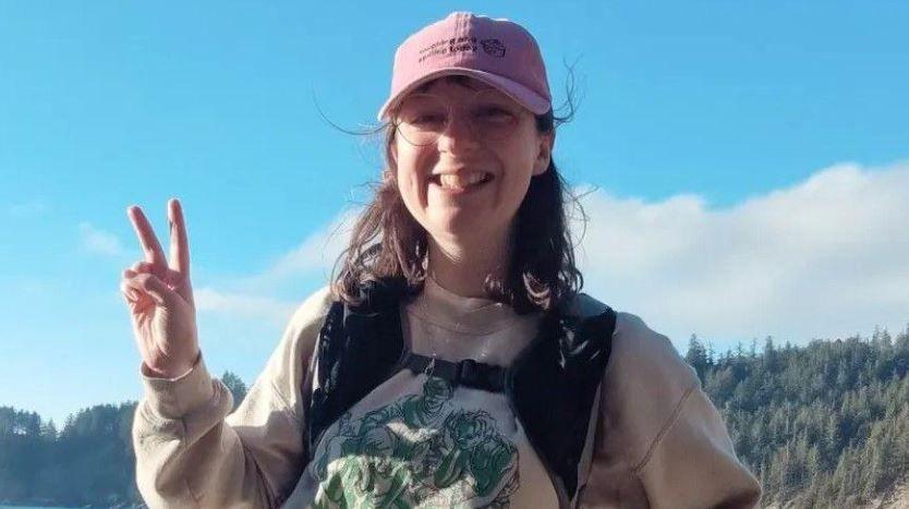 Becky Burke outdoors in front of a forest which can be seen in the far distance. The sun is shining on the left side of her face and she holds her left hand up in a peace sign pose. She smiles widely towards the camera, wearing thin lens round glasses and a pink baseball cap. She has brown short hair and a full fringe peaks out under the cap. 