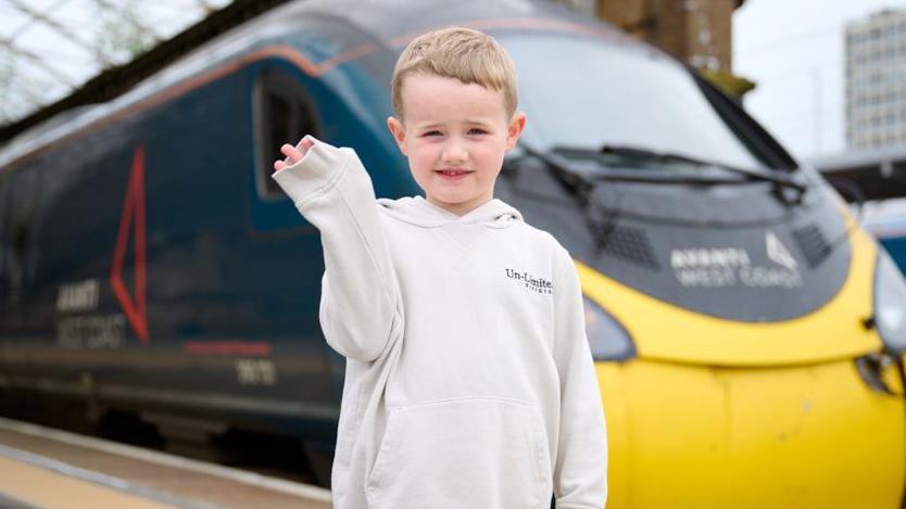 Daniel waves in front of an Avanti train. He is standing on a station platform with the teal and yellow train behind him. Daniel is wearing a cream jumper and waving at the camera.