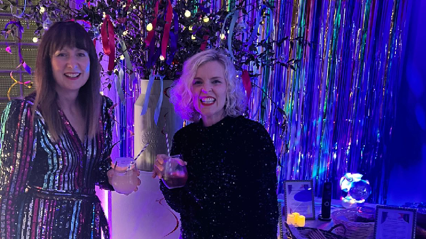 Georgina Jones and Leah Sian Davies at a disco event holding a drink. They are both wearing sparkly outfits. Georgina has long dark hair with a fringe while Leah has blonde chin-length wavy hair and is wearing red lipstick. There are metallic fringe curtain in the background.