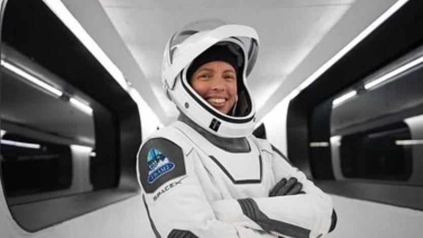 A woman in a white astronaut suit and helmet smiles at the camera with her arms crossed over her chest. The helmet is up so you can see her face. She stands in a white corridor with black windows.