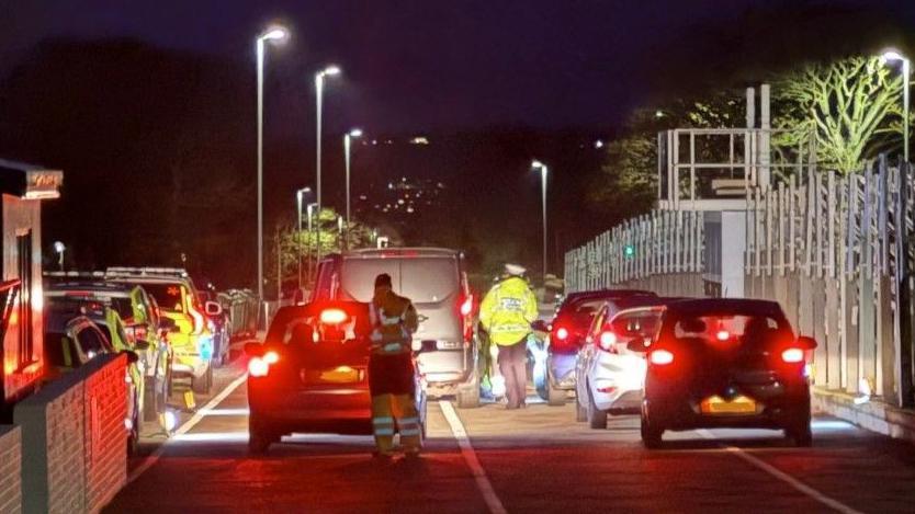 Cars line up in pit lane at the grandstand at night with the rear lights illuminated. There are officers in hi-visibility jackets walking between them