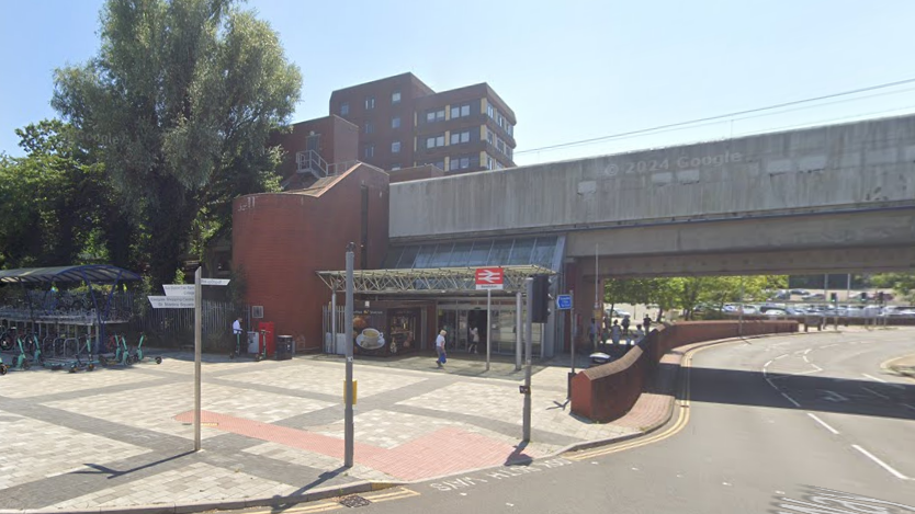 A street view of Basildon Train station. The entrance is under a road bridge, with a train station sign outside. 