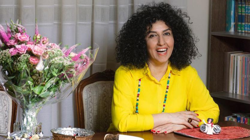 A smiling Narges Mohammadi, wearing a yellow outfit, sits at a table which has a bouquet of flowers on it in 2021