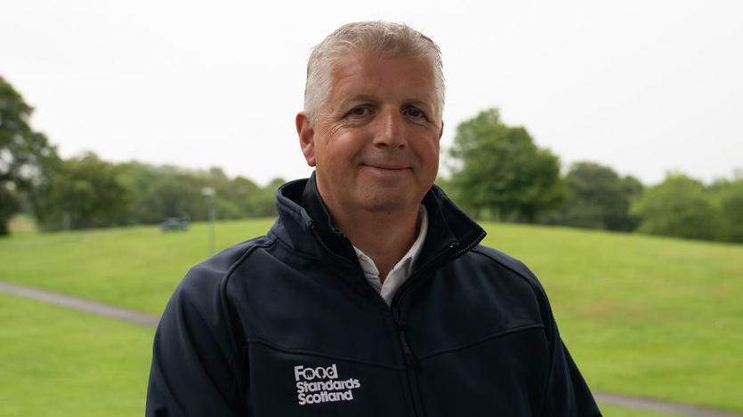 A man with white hair wearing a black jacket with "Food Standards Scotland" emblazoned on it smiles at the camera. He is standing in an open green space like a park