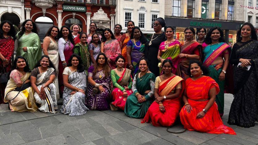 A large group of women posing for the camera in a town square. They wear brightly-coloured sarees.