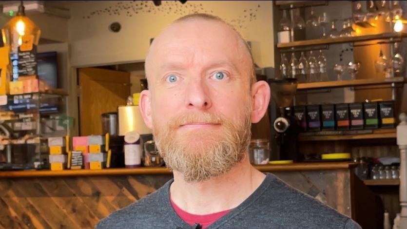 A man with very short strawberry blonde hair and beard and blue eyes looks into the camera. He is standing in a cafe in front of a bar. 