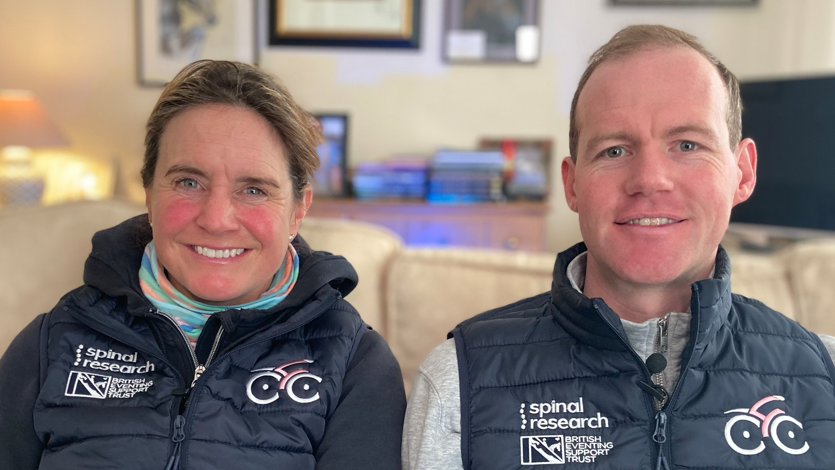 Piggy and Tom March sitting side by side on a cream sofa, both wearing dark blue gilets with Spinal Research and British Eventing Support Trust logos on one side of the chest, and a cycling logo on the other. A table lamp, sideboard, framed pictures on the wall and a TV can be seen behind them. 