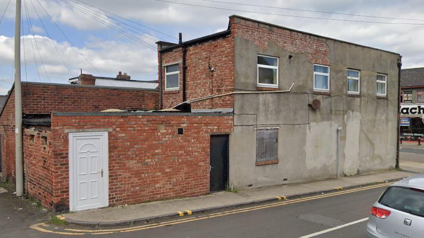 Street view of 8 Stockton Terrace. It is a two-storey building of red brick and peeling cracked grey render. A white door is in the brick wall and upstairs are four windows.