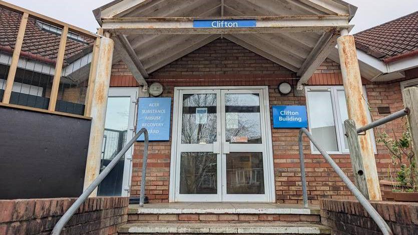 The outside entrance to the detox centre with blue signs that say "Clifton" and "Clifton Building" and "Acer Substance Misuse Recovery Ward"