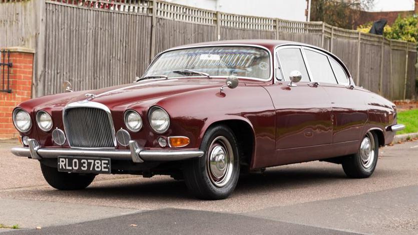 A maroon Jaguar car parked on tarmac with a wooden fence behind it