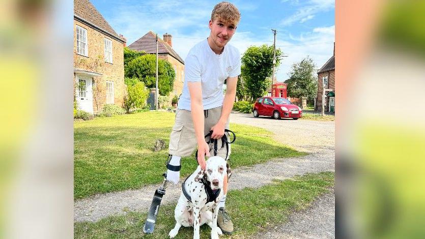 Ollie standing with his prosthetic blade alongside a Dalmatian dog on a lead in a village setting on a sunny day