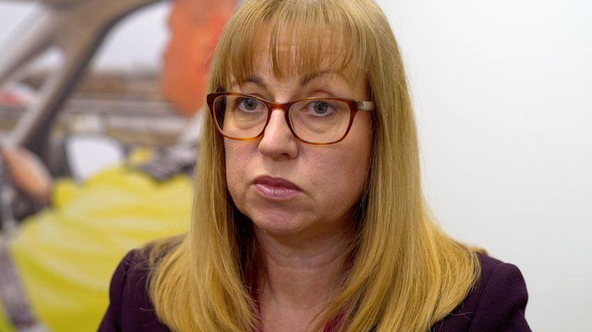 Marian Kitson, who has long, light brown hair and is wearing tortoise shell glasses. She has a serious expression on her face and is standing in front of a picture of a driver wearing a hi-vis jacket.