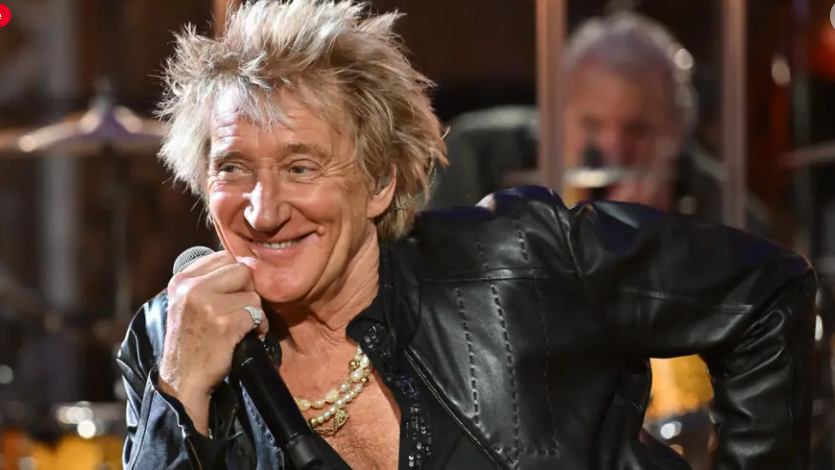 A close up of Rod Stewart in a leather jacket and holding a microphone, smiling at someone to the right of camera