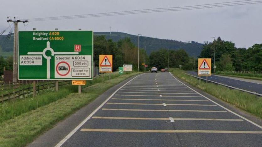 A629 dual carriageway which separates the villages of Steeton and Silsden in Bradford. Fields to the left - hills beyond, road sign with roundabout ahead, road with yellow lines to slow for roundabout, telegraph wires across the road.