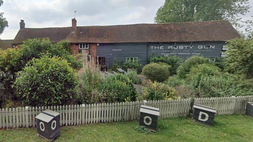 Single-storey grey pub building with "The Rusty Gun" on the back wall in white lettering with a grey background. There is a low wooden fence in the foreground with bushes behind it. There are bee hives in front of the fence.