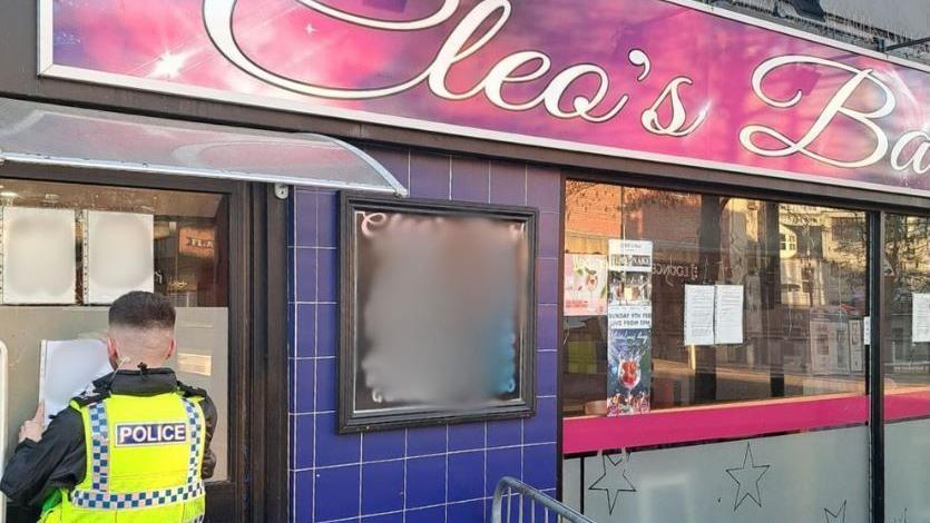 A neighbourhood police officer in a yellow hi-vis jacket with his back to the camera is sticking a closure order on a window of the bar. You can see a canopy and the pink sign of Cleo's bar in curvy white writing. 