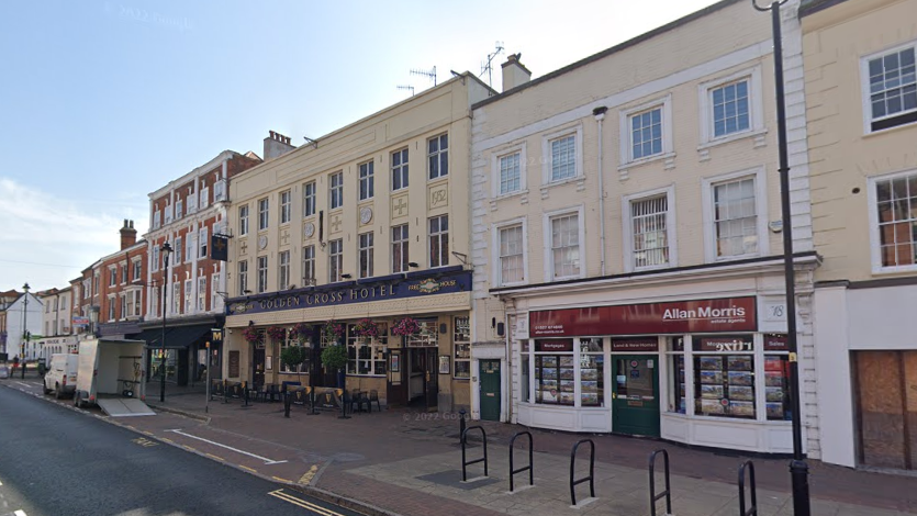 Image taken from the street looking towards the Golden Cross Hotel which is three storeys tall with large square windows. The façade is a pale yellow and above the entrance is a large blue sign with gold writing that reads Golden Cross Hotel. 