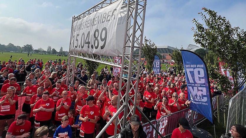 Crowds of runners mainly wearing red running tops waiting to start the Run for the 97 5k in Liverpool's Stanley Park.