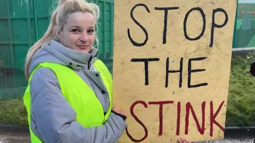 A young woman with long blonde hair, in a grey coat and yellow high vis vest, holds a sign that says 'stop the stink' in front of large green skips at a landfill site.