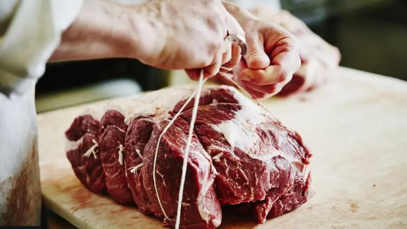 A large chunk of red meat sitting on a butchers block. A person wearing a white coat is tying white string around it.