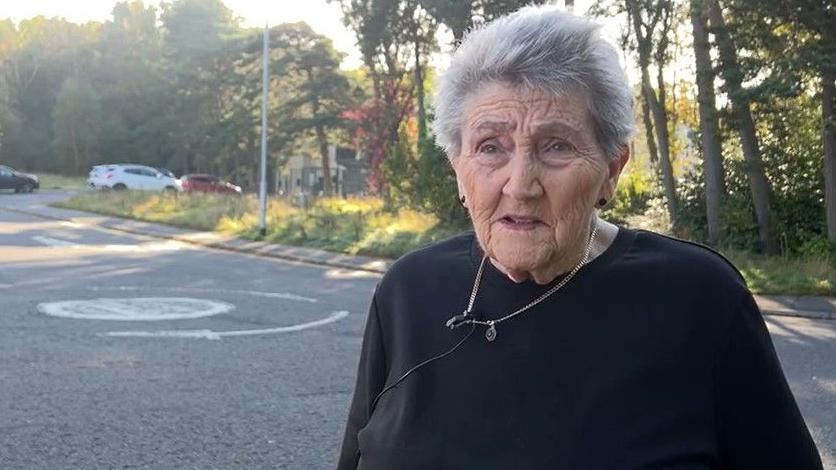 Marion Ward near Shotley Bridge Hospital. She is wearing a black top and has short grey hair. There is a mini roundabout behind her.