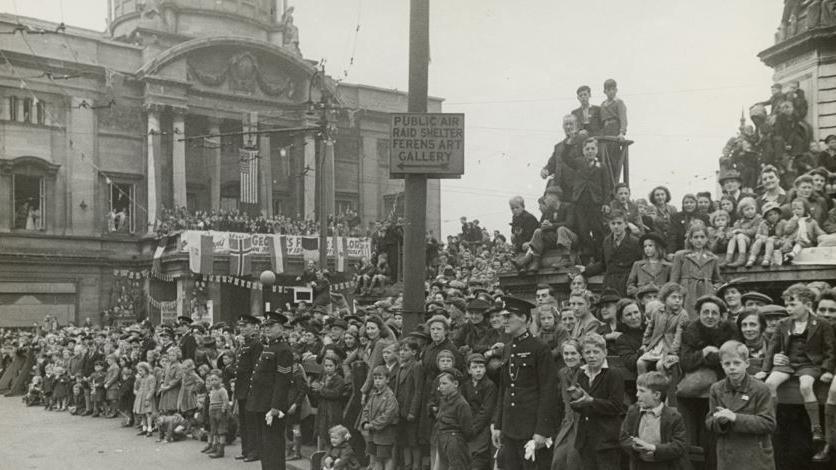 A black and white image, taken in May 1945, showing crowds of people, mostly women and children, in Hull city centre for a victory parade. Some of the children are applauding. Others look solemn.