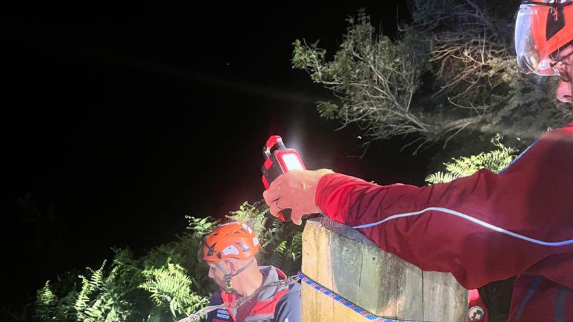 Two rescue team members wearing orange safety helmets with head torches and red rescue clothing. One standing beside a wooden post holding a torch. A blue-and-white rope is around the post. Another rescuer is lower on the slope, also wearing an orange helmet and rescue gear.