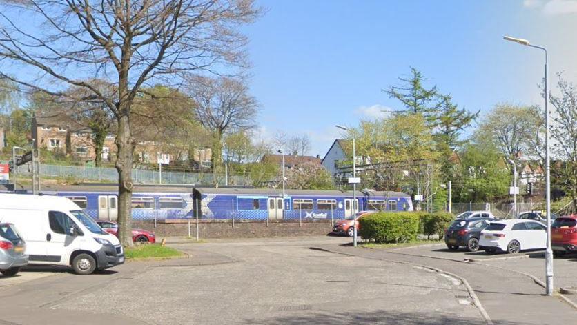 A residential street with cars parked in bays along the side. A ScotRail train can be seen on the tracks next to the street.