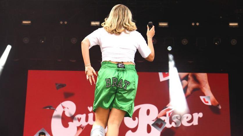 Singer Caity Baser seen from behind on stage, in green silk shorts with the word brat sewn onto them, plus a white T shirt