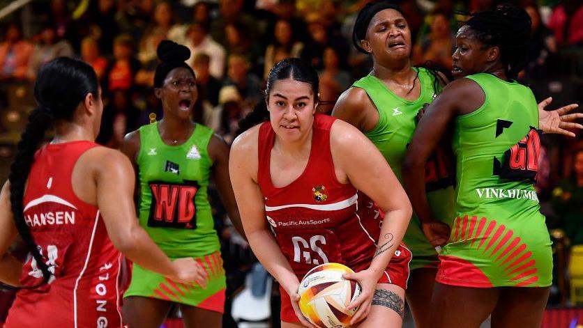 Uneeq Palave, in Tonga's red kit, holds the netball during a match against Malawi at the 2023 World Cup. Malawi players, in green kit, have alarmed expressions on their faces.