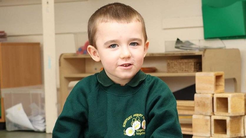 Lincoln Button wearing his school uniform, which is a green sweater that has a green polo top underneath. He has short brown hair and is smiling at the camera while sitting in a classroom. 