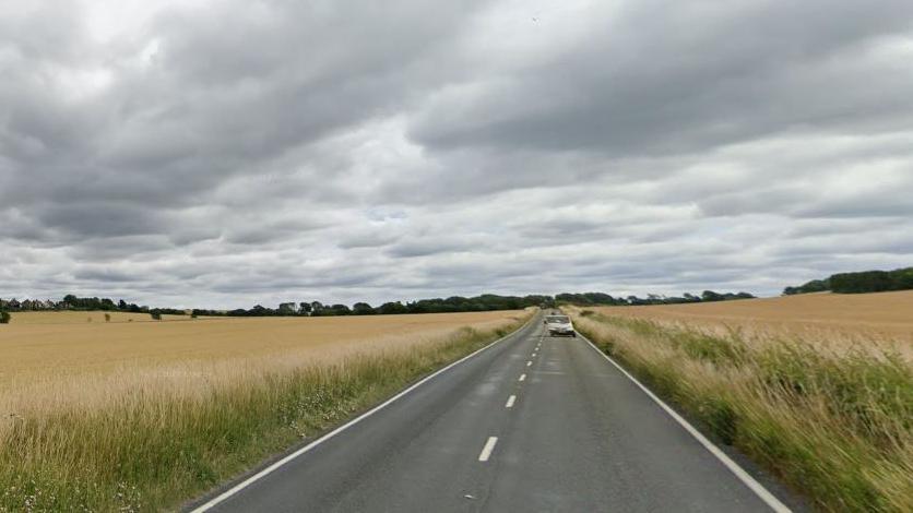 A rural road with crops growing on either side of the road, dual carriageway with a white car in the distance