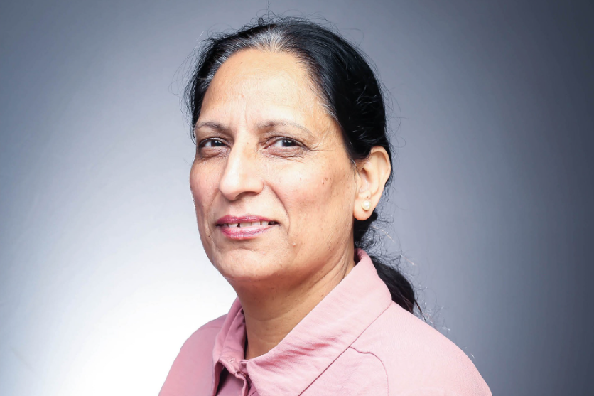 A middle-aged woman with black hair tied back in a ponytail smiles at the camera. She wears a pink shirt and has white pearl-like earrings in. The photo is a professional headshot and she is pictured in front of a grey background.