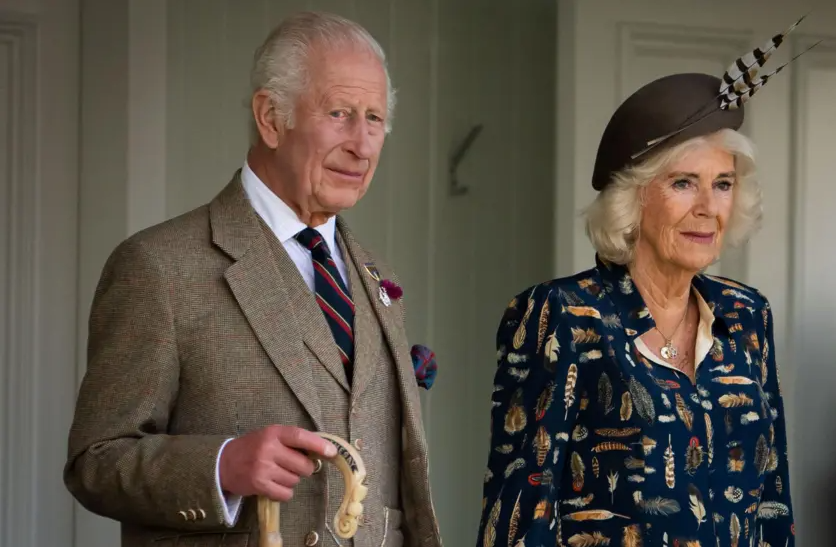 King Charles wearing a tweed suit with shirt and tie holding a shepherd's crook and standing next to Queen Camilla who is wearing a blue patterned dress and a blue hat with two feathers attached