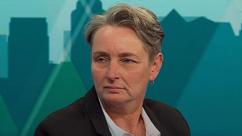 A woman with short hair in a black jacket and white shirt sits on a sofa in a TV studio