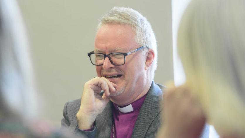 The Bishop of Grimsby, wearing a tweed jacket and traditional purple shirt, talks to two people who have their backs turned to the camera. He is smiling and has his hand on his chin.