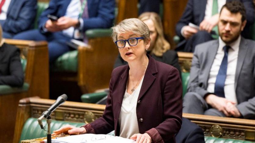 Yvette Cooper standing at the dispatch box in the House of Commons. She is wearing a maroon blazer, white blouse, and blue glasses. The benches behind her are filled with MPs.