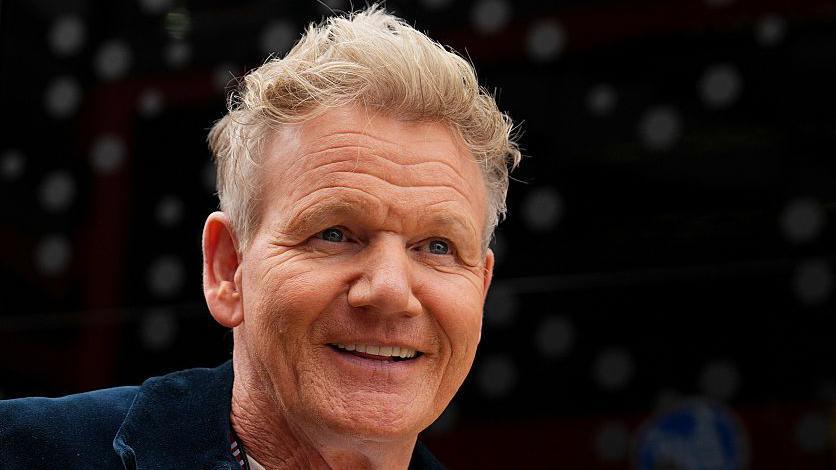 Gordon Ramsay, a man with short blonde hair wearing a black zip-up jacket over a white t-shirt. He is smiling in front of a blurred black background.