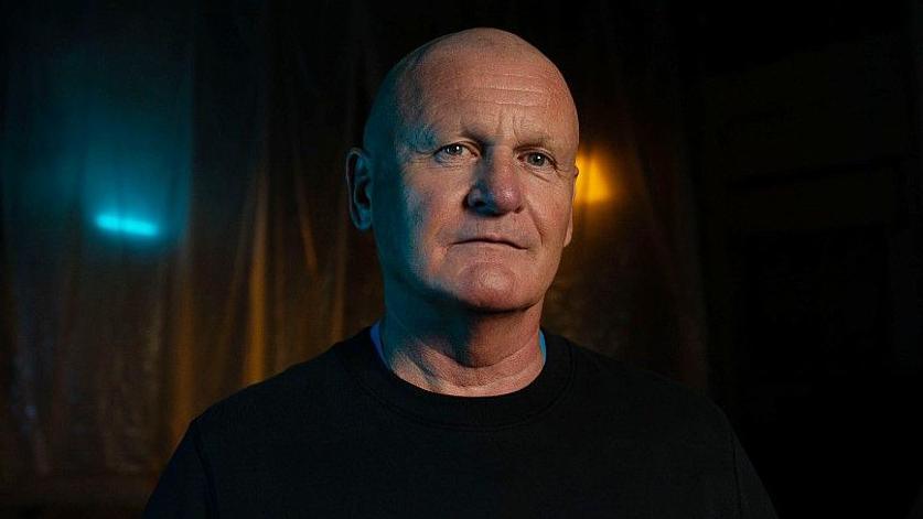 Andrew Dodd has a bald head and blue eyes and is lightly tanned. He is approximately in his late 50s. He is looking at the camera. He wears a plain black jumper. The background is dimly lit with lights behind a translucent curtain. It is a head and shoulders shot of him. 