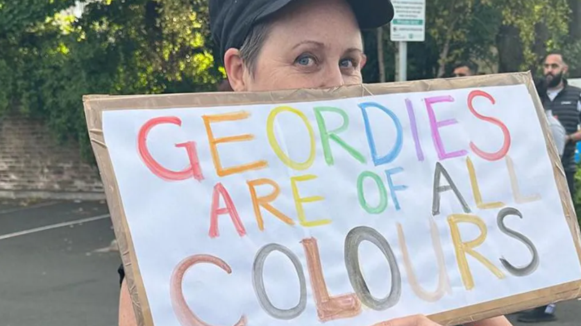 A woman with short hair in a black cap holding a sign which reads 'Geordies are of all colours' written with multi-coloured letters.