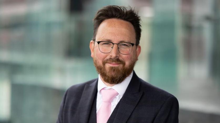 A man with dark hair, glasses and a beard wearing a suit. 