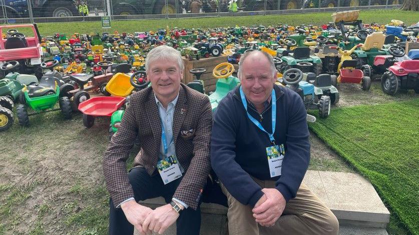 Haydn Evans and Robert Powell sit on the ground in front of a number of toy tractors, with tractors lined up on the road behind them. Both men are smiling at the camera.