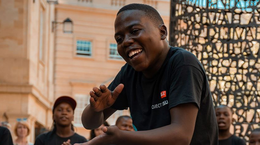 A Project Zulu choir member clapping his hands and smiling as he dances to the music. He's wearing a black t-shirt with 'Project Zulu' above a logo on the right of the top.