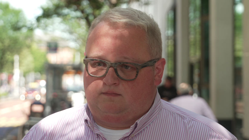 Séamas de Fáoite has short grey hair, a round face and is wearing light grey square glasses. He is wearing a pink and white stripe shirt with a white t-shirt on beneath it.