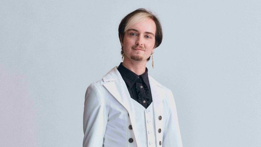 Stanley Baxton wears a white suit jacket over a black shirt. He has dark hair with a blonde fringe. 