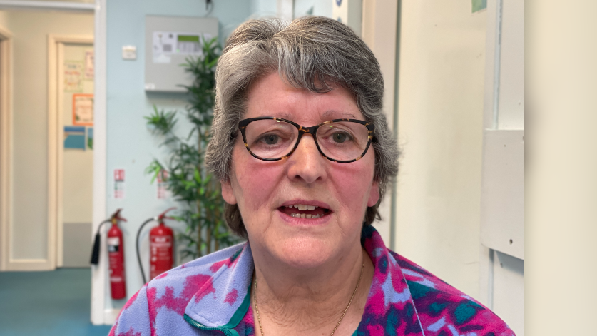 A woman looks at the camera. She has short grey hair and glasses. She is wearing a top patterned with different shades of purple. A corridor with a potted plant is visible in the background. 