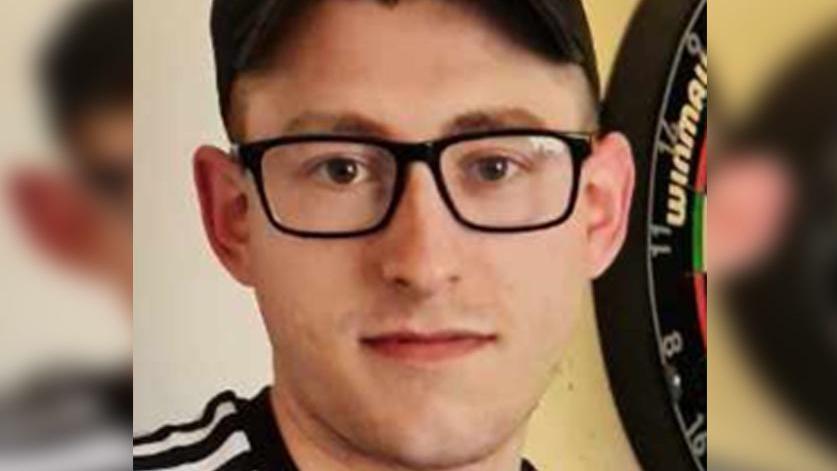 Martyn Richardson, wearing glasses and a cap, looking at the camera. He is standing by a dart board and wearing a black and white top.