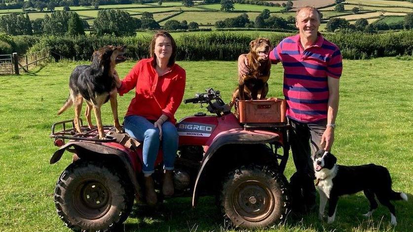 Kirsty Williams, pictured with her husband Richard. They are on a farm. There is grass behind them. She is sat on a red quad bike along with two dogs. He is stood next to the bike and has his arm around one dog and another black and white dog at his feet. He wears jeans and a pink and blue striped top. She has a red top and blue jeans on.